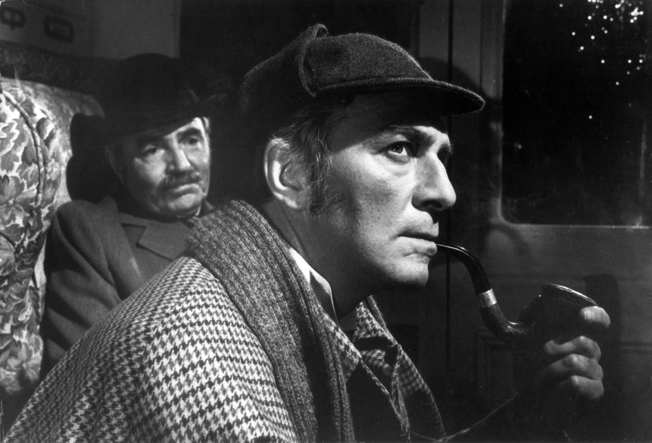 Christopher Plummer als Sherlock Holmes in dem Film "Murder by Decree" (1979). Im Hintergrund James Mason als Dr. Watson. Holmes sitzt in einem Zugabteil und raucht eine Pfeife. Er guckt nachdenklich.
