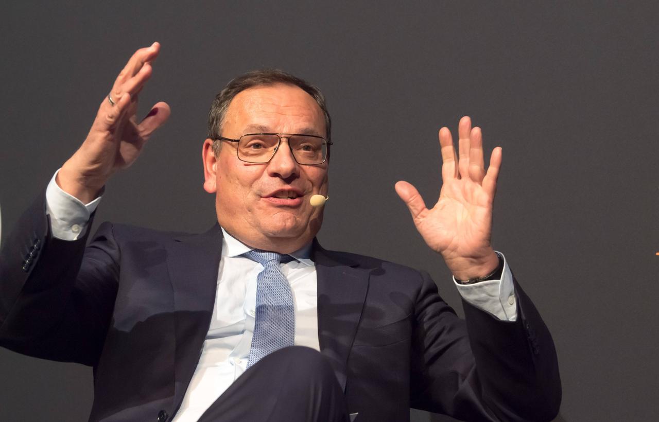Hans-Jürgen Jakobs sitzt auf einer Bühne und hält beide Hände in die Luft, als erkläre er etwas. Hans-Jürgen Jakobs sitzt auf einer Bühne und hält beide Hände in die Luft, als erkläre er etwas.