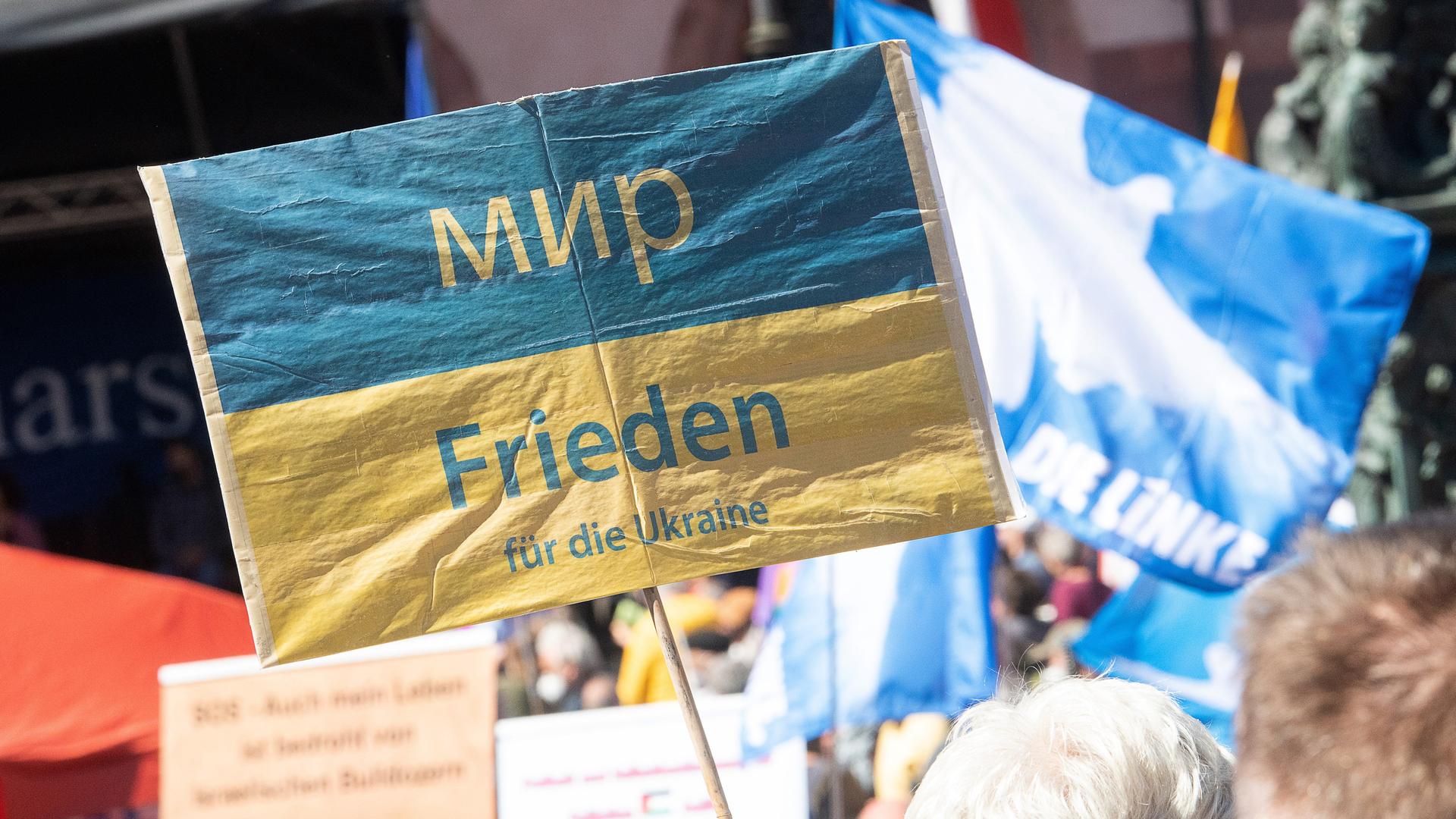 «Frieden für die Ukraine» steht während der Abschlusskundgebung der Ostermärsche des «Netzwerks Friedenskooperative» in Frankfurt und Umgebung auf einem Schild. Im Hintergrund ist eine Fahne mit Friedenstaube zu sehen. 