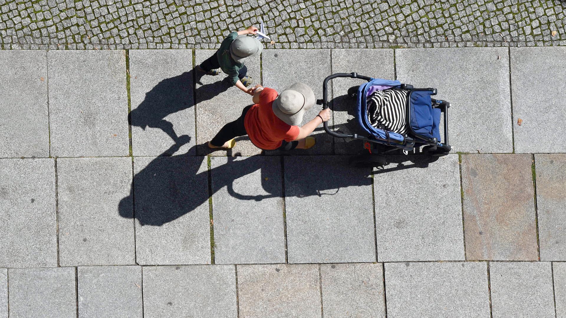 Mit Hut und Mütze als Sonnenschutz gehen eine Frau mit Kinderwagen und ein Kind einen Gehweg entlang.