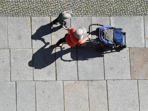 Mit Hut und Mütze als Sonnenschutz gehen eine Frau mit Kinderwagen und ein Kind einen Gehweg entlang.