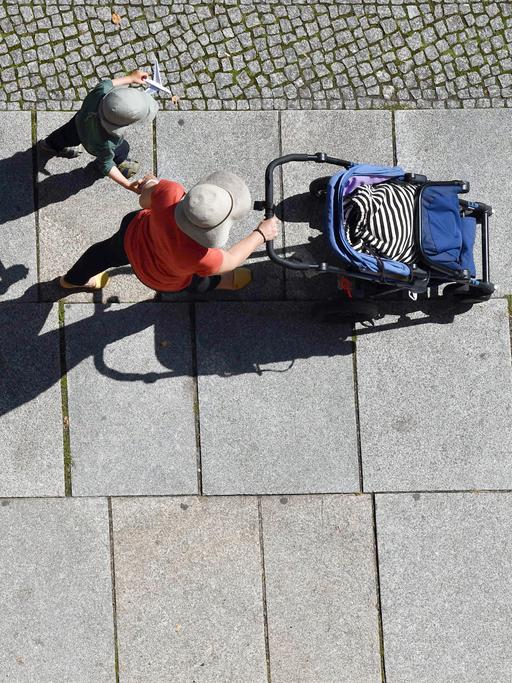 Mit Hut und Mütze als Sonnenschutz gehen eine Frau mit Kinderwagen und ein Kind einen Gehweg entlang.