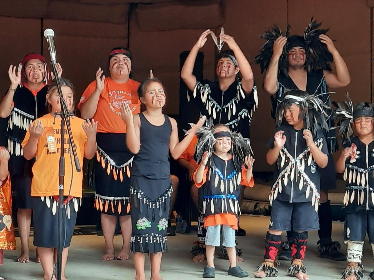 Indigene Frauen und Kinder stehen und tanzen auf einer Bühne, mit Federschmuck und tradioneller Kleidung. Aufführung der Penelakut Drummers and Dancers während der Gedenkveranstaltung in Chemainus