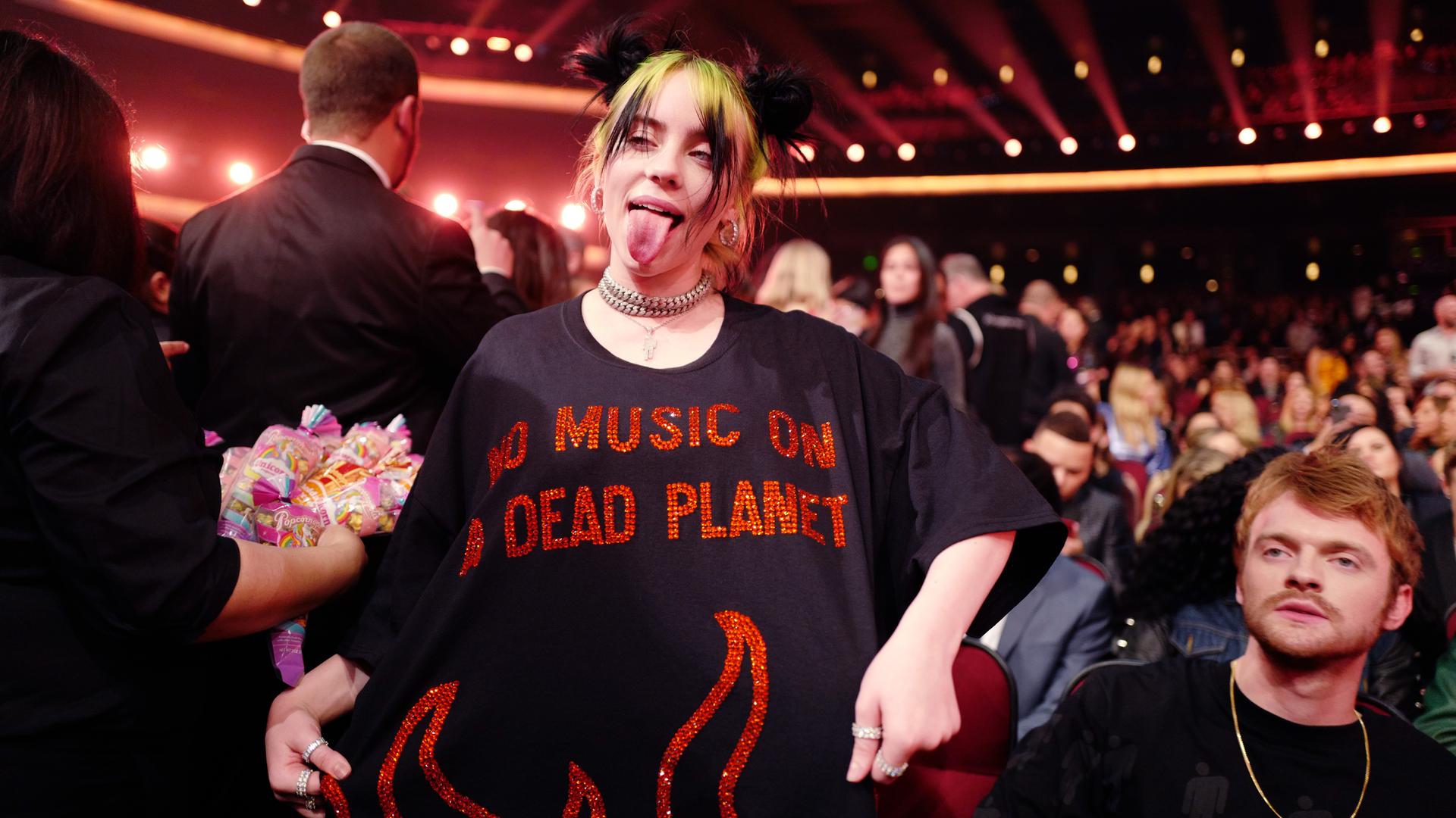 Billie Eilish streckt die Zunge heraus und zeigt ihr T-Shirt vor, auf dem in roten, pailettenbesetzten Buchstaben steht: "No Music On A Dead Planet". Billie Eilish streckt die Zunge heraus und zeigt ihr T-Shirt vor, auf dem in roten, pailettenbesetzten Buchstaben steht: "No Music On A Dead Planet".