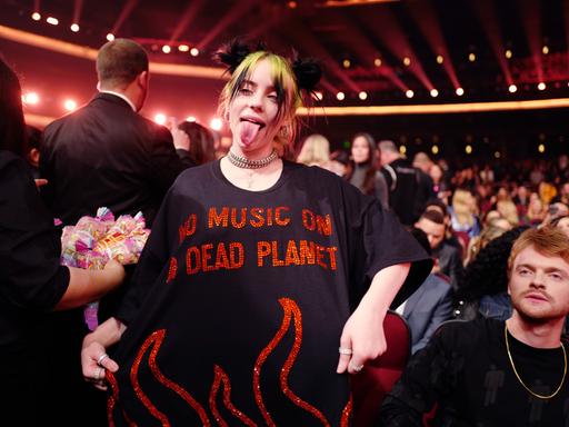 Billie Eilish streckt die Zunge heraus und zeigt ihr T-Shirt vor, auf dem in roten, pailettenbesetzten Buchstaben steht: "No Music On A Dead Planet".
