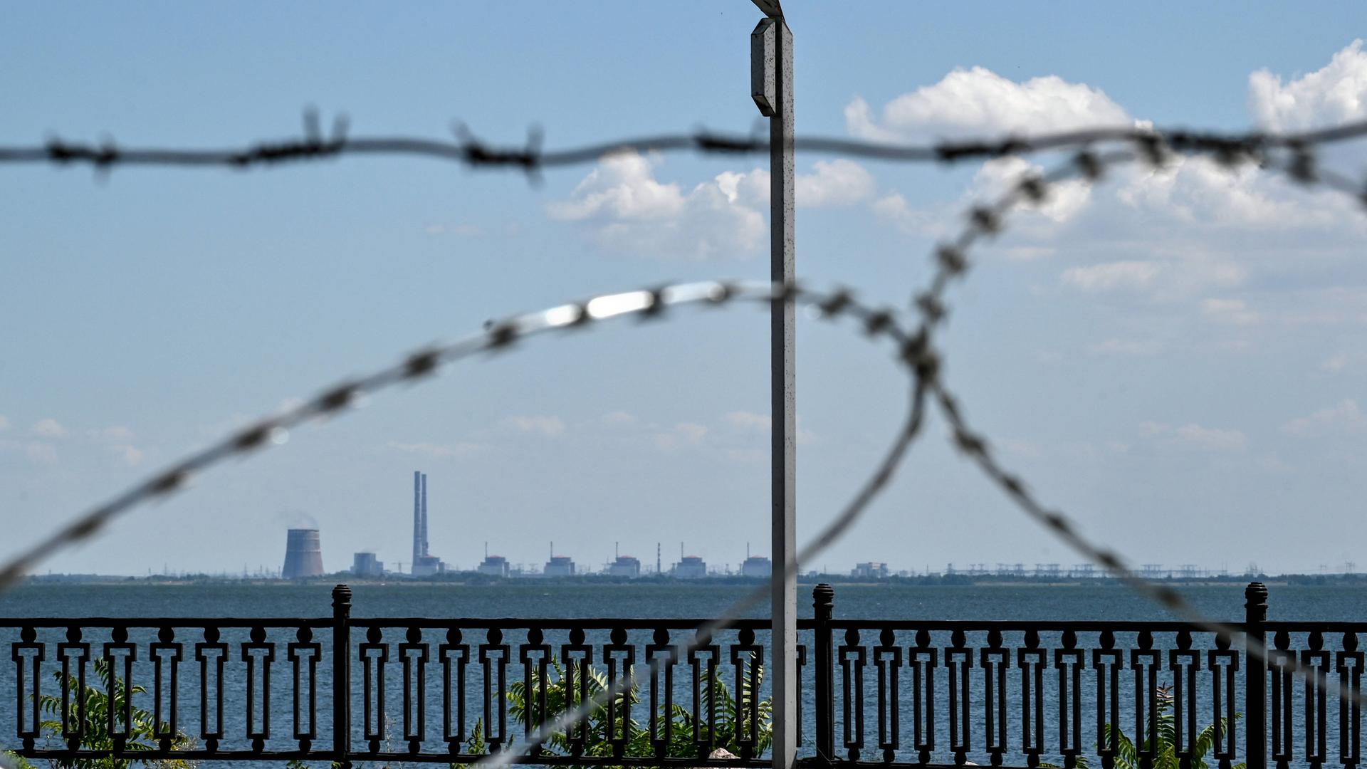 Blick durch Stacheldraht und über einen hohen Zaun auf das ukrainische Atomkraftwerk Saporischschja, das in der Ferne am Horizont zu sehen ist. Blick durch Stacheldraht und über einen hohen Zaun auf das ukrainische Atomkraftwerk Saporischschja, das in der Ferne am Horizont zu sehen ist.