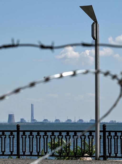 Blick durch Stacheldraht und über einen hohen Zaun auf das ukrainische Atomkraftwerk Saporischschja, das in der Ferne am Horizont zu sehen ist.
