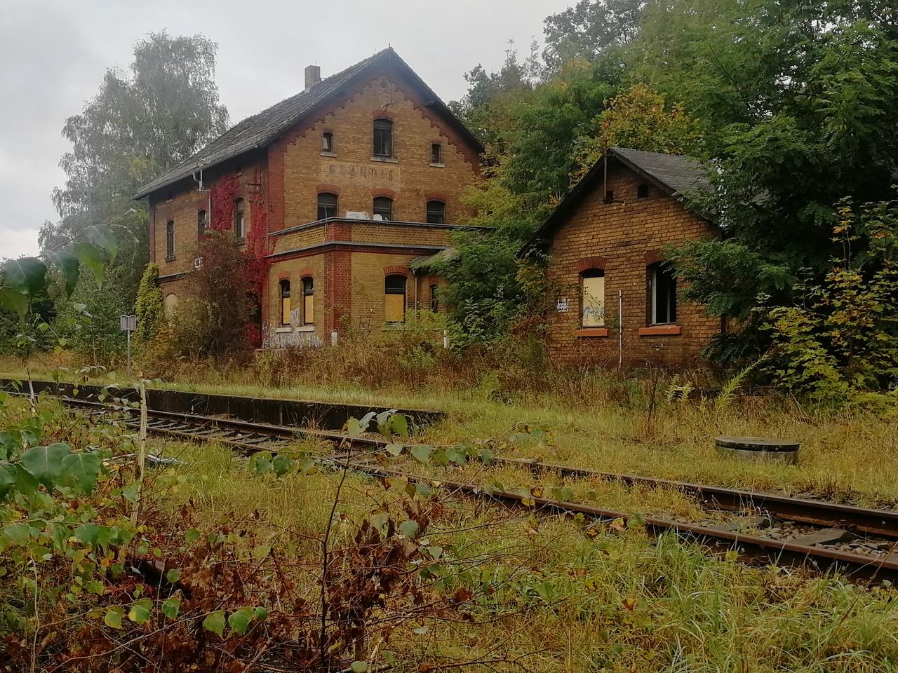 Ein altes Bahnhofsgebäude aus gelben Ziegelsteinen. Die Fenster sind kaputt oder zugemauert. Auf den Gleisen davor wachsen Gräser.