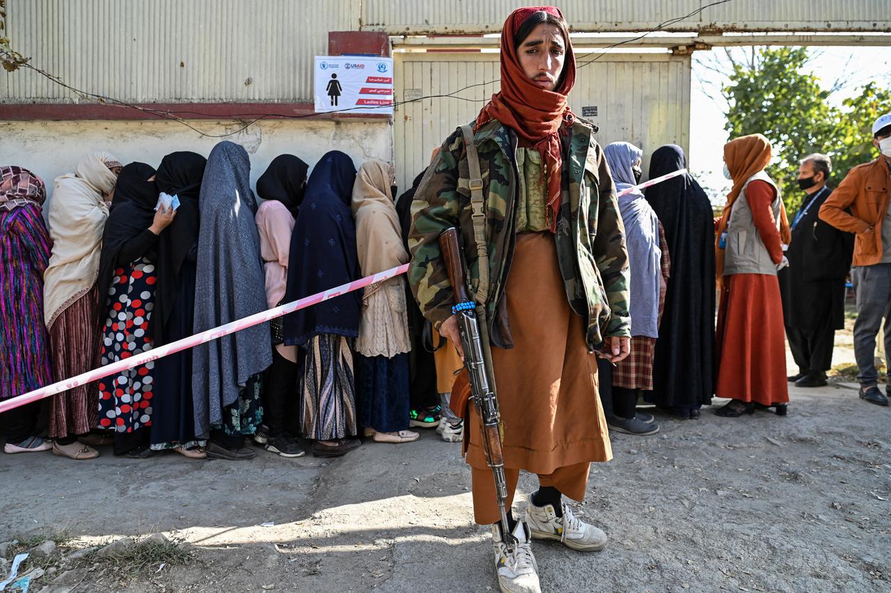 Ein bewaffneter Talibankämpfer schaut in die Kamera, während hinter ihm verschleierte Frauen an einer Essensausgabe anstehen.