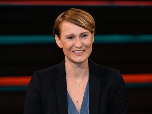 Jana Puglierin trägt ein schwarzes Sacko, eine blaue Bluse und sitzt vor einem Studiohintergrund. Sie schaut lächelnd in die Kamera.