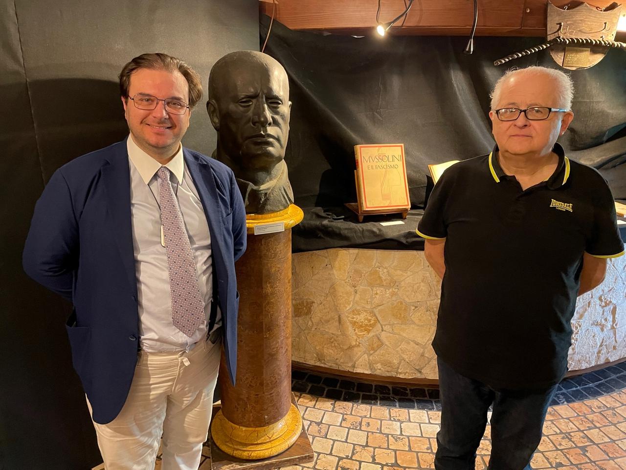 Ein jüngerer Herr mit Anzug und Krawatte steht gemeinsam mit einem älteren Herrn strahlend neben einer großen schwarzen Mussolini-Büste. Ein jüngerer Herr mit Anzug und Krawatte steht gemeinsam mit einem älteren Herrn strahlend neben einer großen schwarzen Mussolini-Büste.
