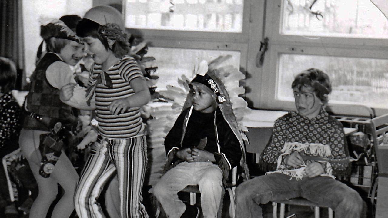Verkleidete weiße Kinder feiern Fasching in einem Klassenzimmer. Zwei Kinder tanzen, ein schwarzer Junge im Indianerkostüm sitzt traurig auf einem Stuhl daneben. Verkleidete weiße Kinder feiern Fasching in einem Klassenzimmer. Zwei Kinder tanzen, ein schwarzer Junge im Indianerkostüm sitzt traurig auf einem Stuhl daneben.
