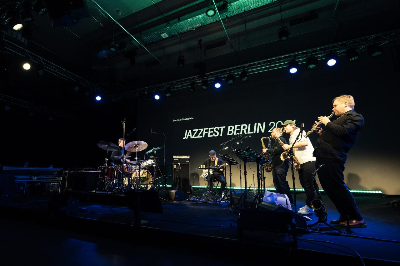 Eine Band aus, von links, Schlagzeuger, E-Bassist und drei Saxofonisten spielt auf einer Bühne. Im Hintergrund der Schriftzug "Jazzfest Berlin".