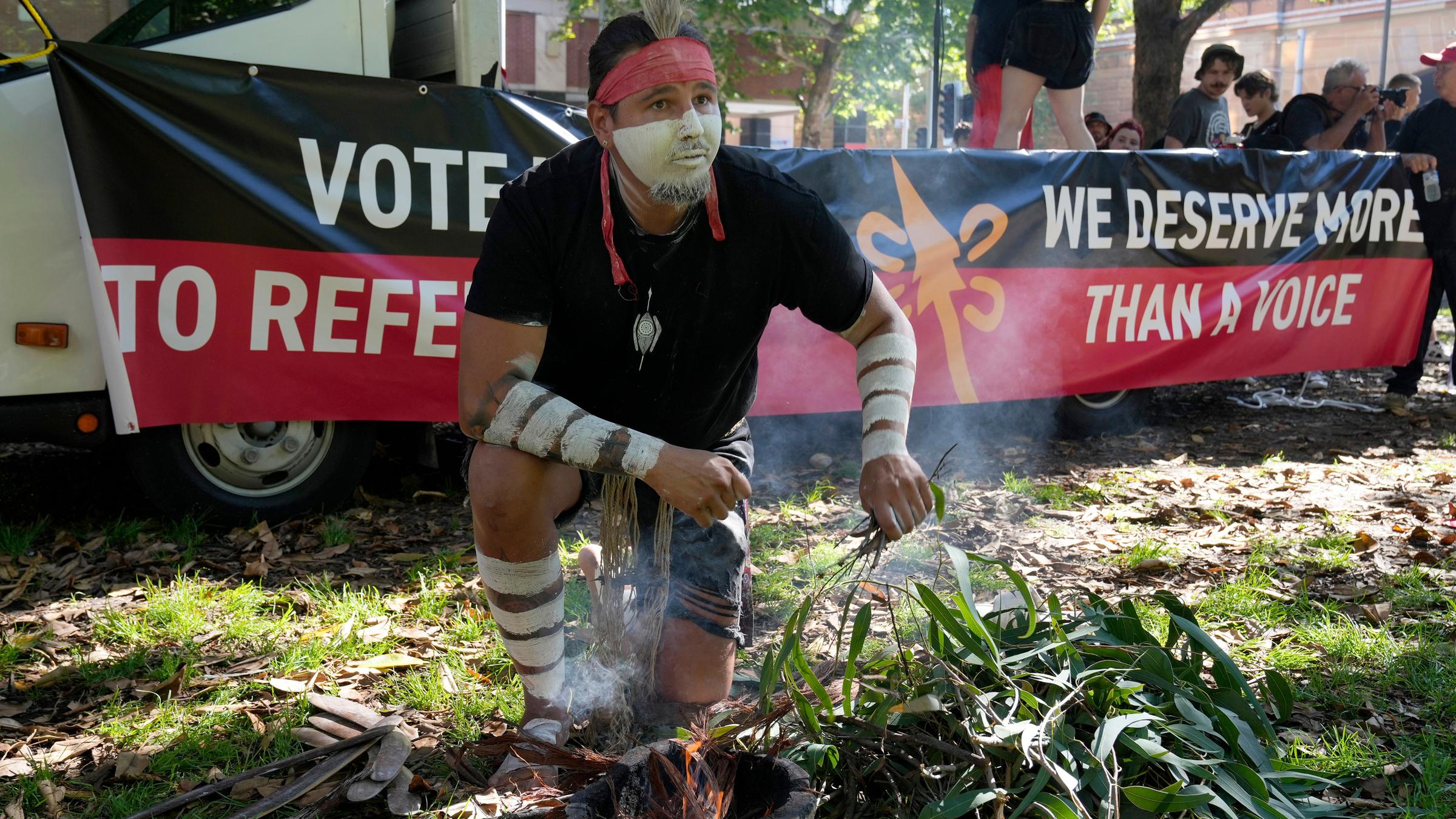 Ein Aboriginal mit weißer Gesichtsbemalung entfacht ein zeremonielles Feuer. Hinter ihm steht auf einem Spruchband: "We deserve more than a voice." 