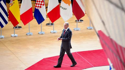 Bundeskanzler Olaf Scholz im Gebäude des Europäischen Rates in Brüssel. Er geht auf einem roten Teppich, vorbei an den Flaggen der Mitgliedsländer der EU.
