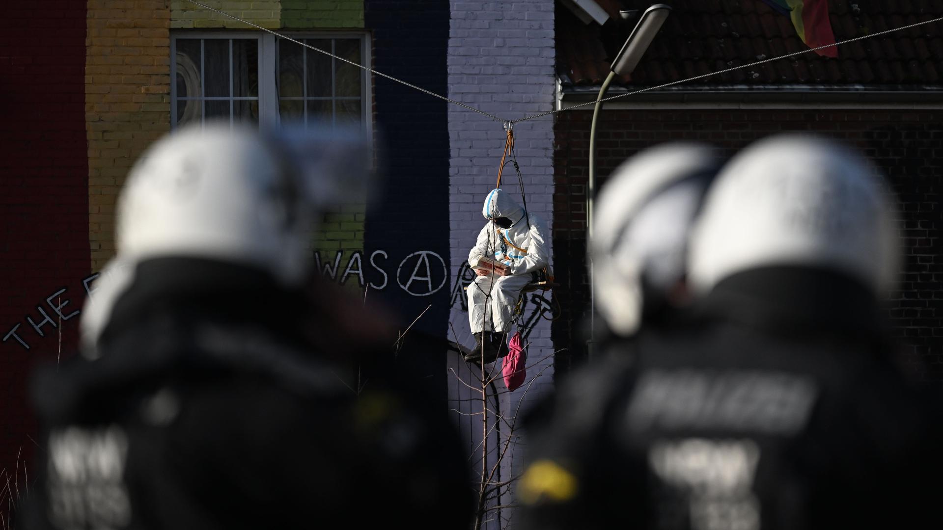 Ein Demonstrant sitzt während der Räumung des Dorfes Lützerath auf einem Brett, das an einem Drahtseil hängt. Polizisten beobachten die Person. Ein Demonstrant sitzt während der Räumung des Dorfes Lützerath auf einem Brett, das an einem Drahtseil hängt. Polizisten beobachten die Person.