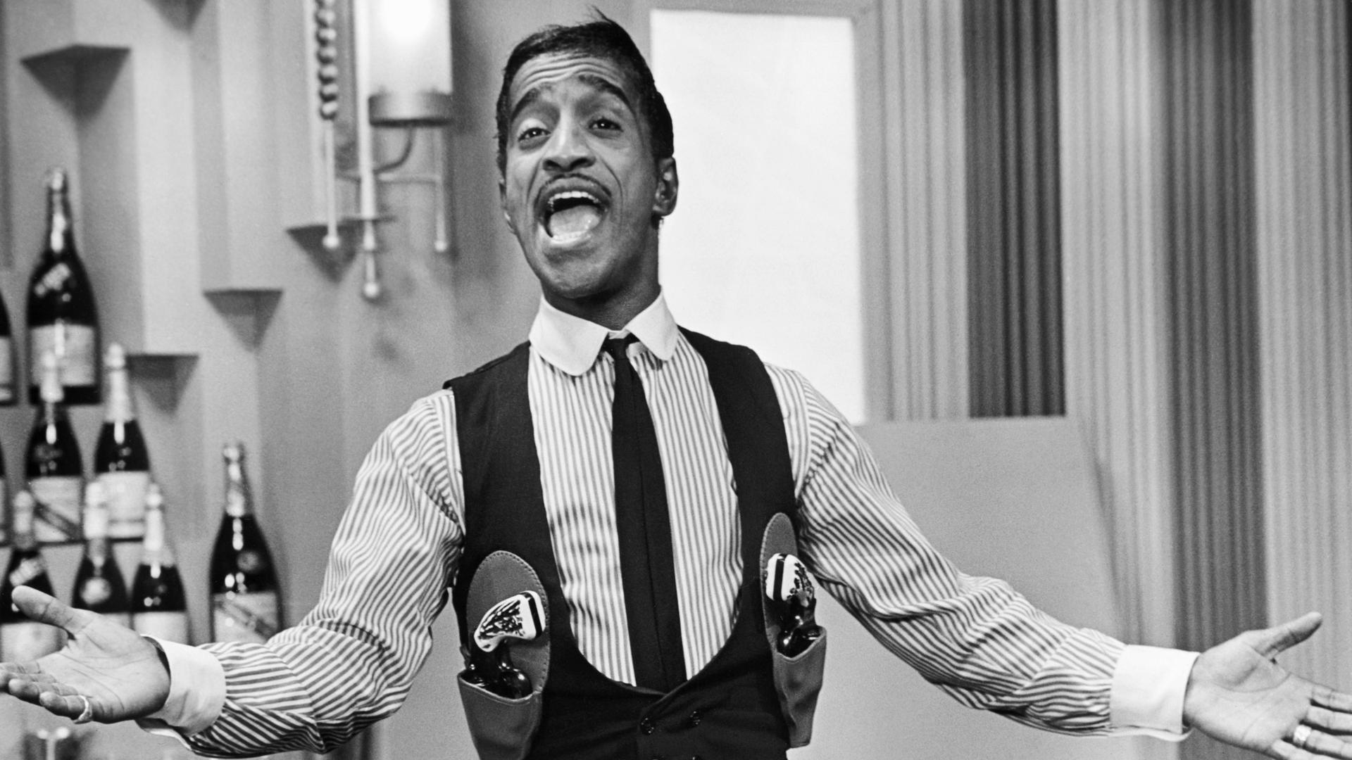 Schwarz-weiss Porträt von Sammy Davis Jr. mit Pistolengurt. Er singt und hat die Arme ausgebreitet. Schwarz-weiss Porträt von Sammy Davis Jr. mit Pistolengurt. Er singt und hat die Arme ausgebreitet.