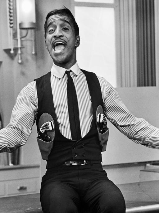 Schwarz-weiss Porträt von Sammy Davis Jr. mit Pistolengurt. Er singt und hat die Arme ausgebreitet.