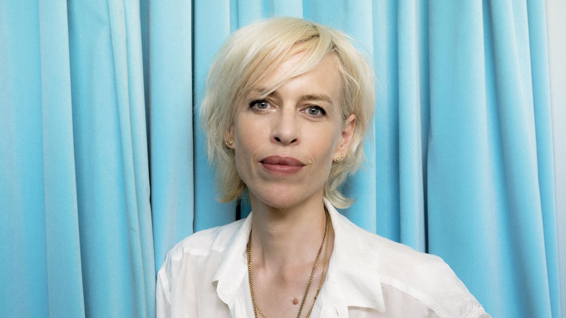 Porträtfoto der Journalistin und Autorin Katja Eichinger. Sie trägt eine weiße Bluse und steht vor einem hellblauen Vorhang.