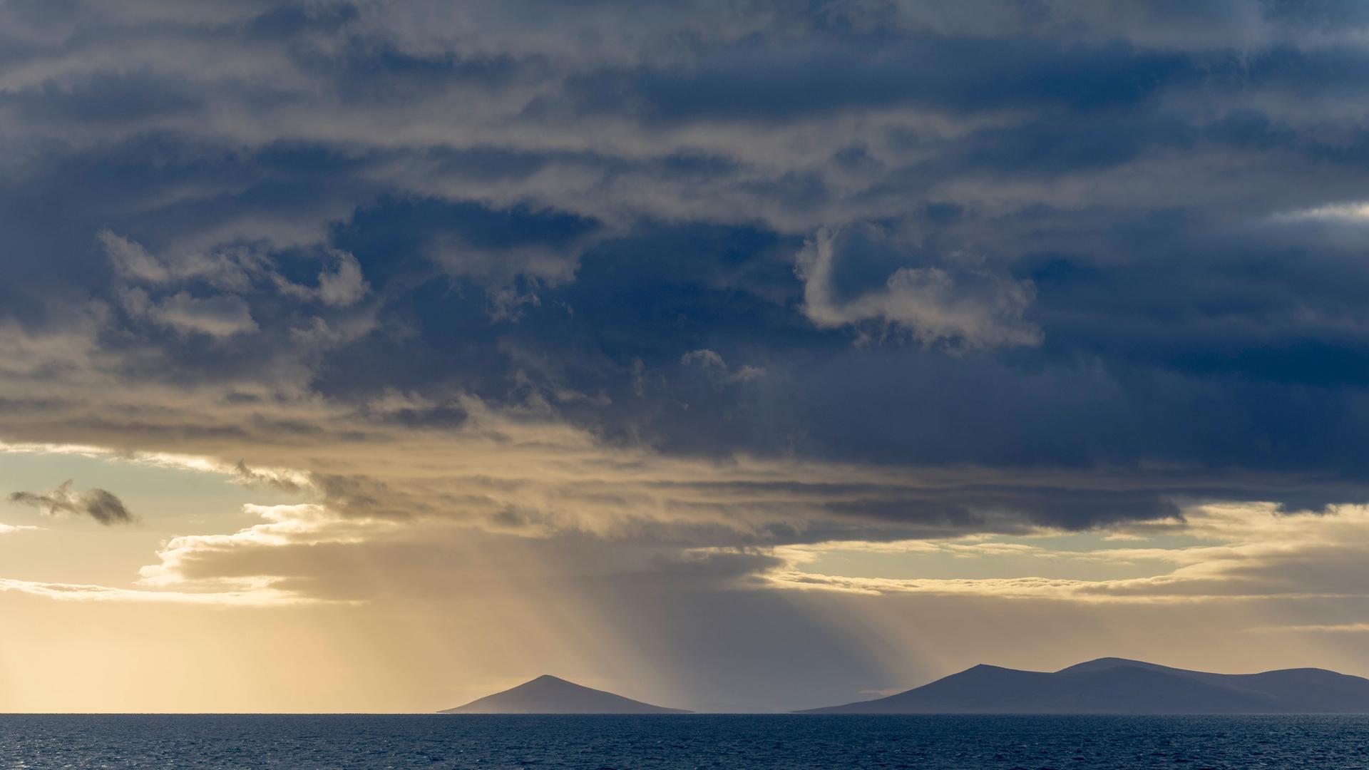 Stürmische dunkle und helle Wolken über flachen Inselkuppen im Meer mit einem Hauch von Sonne.