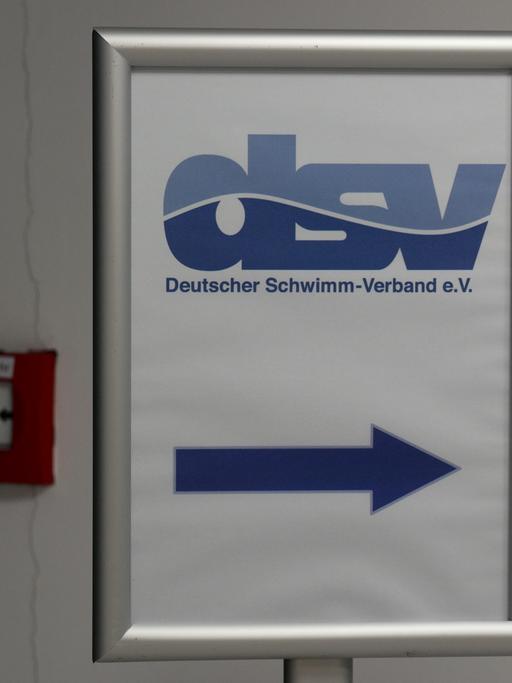 Auf dem Schild mit blauer Schrift auf weißem Grund zeigt ein pfeil nach rechts. Es hängt an einer Wand neben einem Feuer-Notmeldeknopf. 