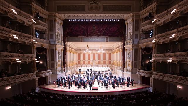 Ein großer Opernsaal ist zu sehen, mit einer tiefen Bühne und hohen Zuschauerrängen. Auf der Bühne steht ein Orchester. Es ist die Sächsische Staatskapelle in der Semperoper in Dresden.