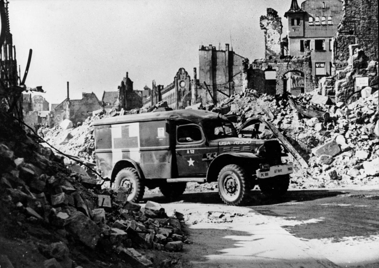 Ein Krankenwagen fährt in diesem Schwarz-Weiß-Filmstill durch eine Trümmerlandschaft.