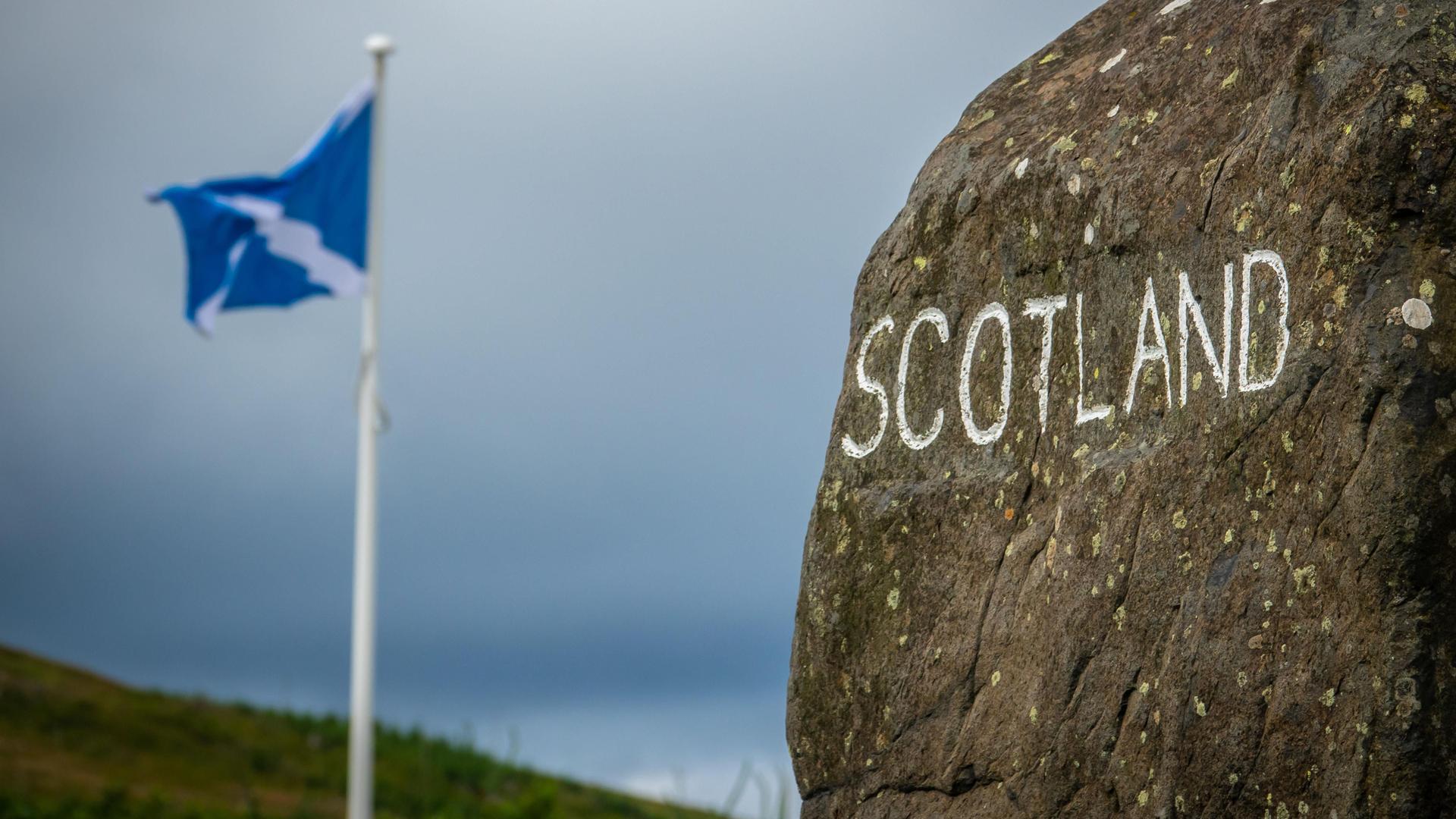 Auf einem Stein steht mit weißer Farbe geschrieben "Schottland". Dahinter weht eine schottische Fahne. Auf einem Stein steht mit weißer Farbe geschrieben "Schottland". Dahinter weht eine schottische Fahne.