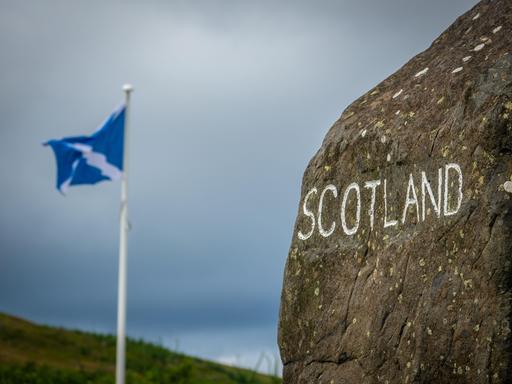 Auf einem Stein steht mit weißer Farbe geschrieben "Schottland". Dahinter weht eine schottische Fahne.