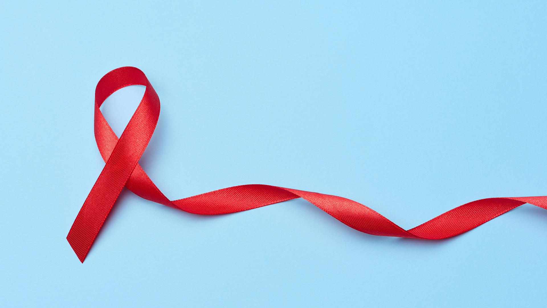 Eine rote Aids-Solidaritätsschleife, deren eines Ende länger ist und in der Spiralform einer DNA zur Seite fließt.