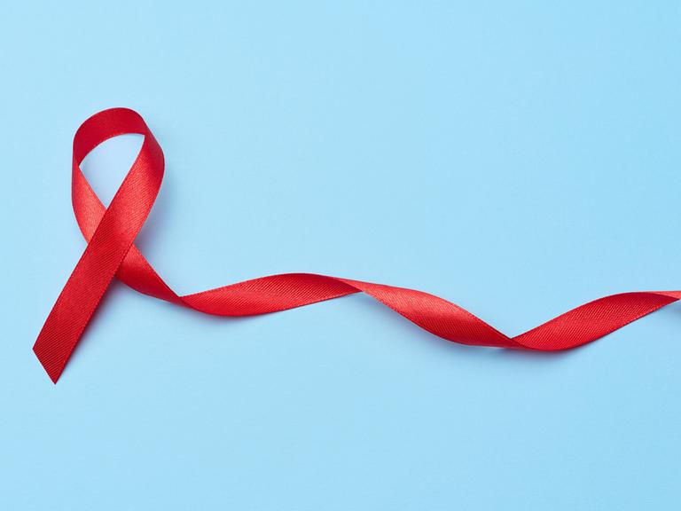 Eine rote Aids-Solidaritätsschleife, deren eines Ende länger ist und in der Spiralform einer DNA zur Seite fließt.