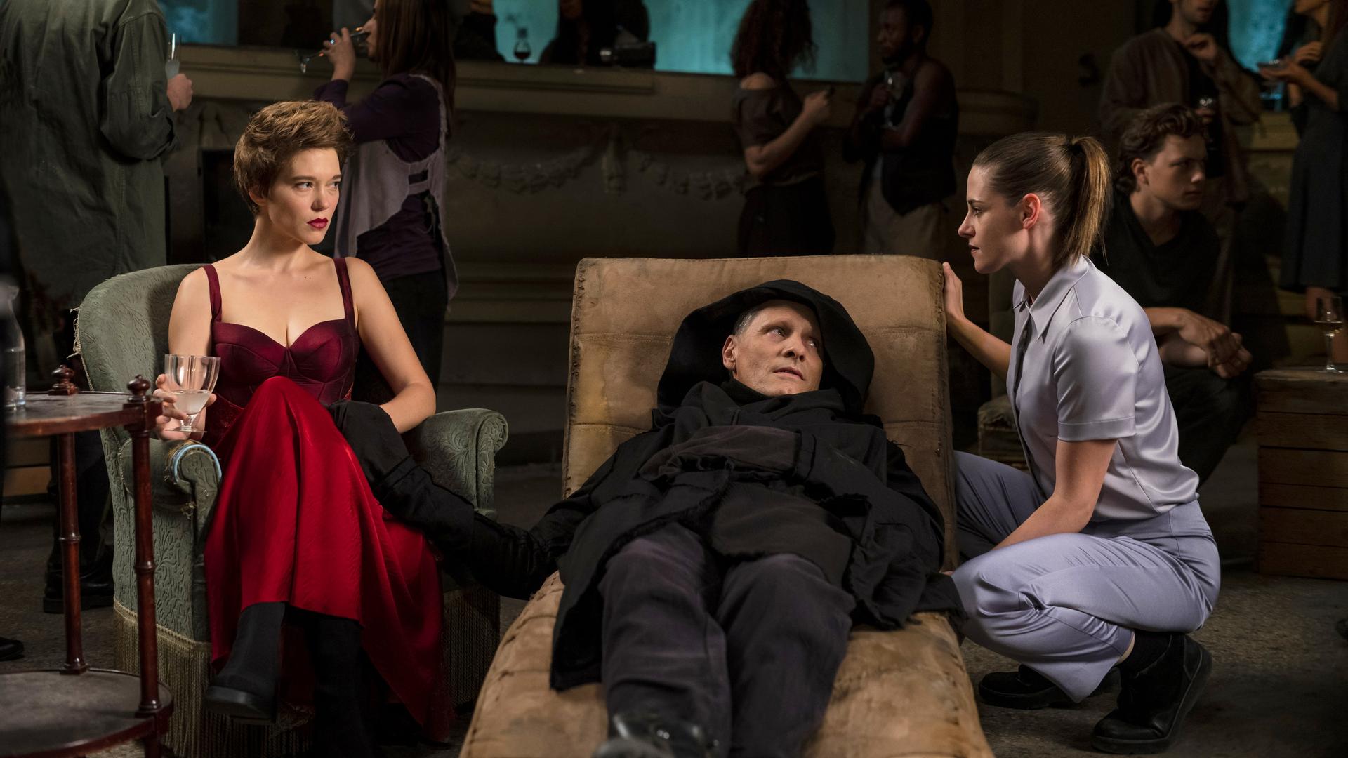 Ein Mann (Viggo Mortensen) liegt auf einer Liege, neben ihm sitzt auf einem Sessel eine Frau (Léa Seydoux), eine weitere (Kristen Stewart) ist neben der Liege in die Hocke gegangen.