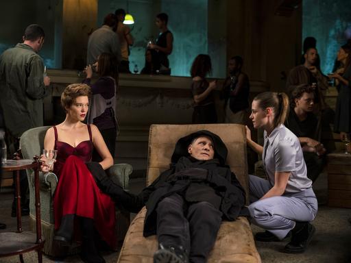 Ein Mann (Viggo Mortensen) liegt auf einer Liege, neben ihm sitzt auf einem Sessel eine Frau (Léa Seydoux), eine weitere (Kristen Stewart) ist neben der Liege in die Hocke gegangen.