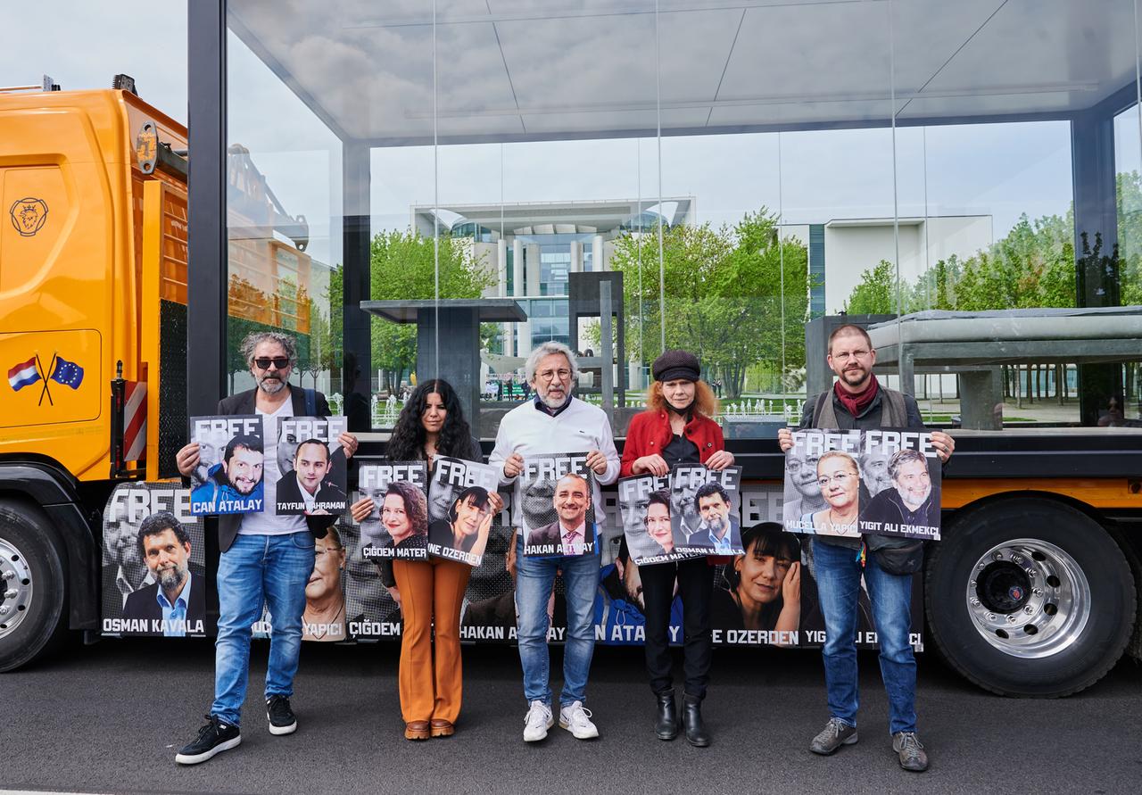 Protest in Berlin gegen die Verurteilung von Kavala: Die ehemals politischen Gefangenen in der Türkei (von links nach rechts) Deniz Yücel, Zehra Dogan, Can Dündar, Asli Erdogan und Peter Steudtner stehen vor dem Bundeskanzleramt vor einem Nachbau einer Zelle. Sie fordern die Freilassung weiterer Menschenrechtsaktivisten, welche auf den Plakaten zu sehen sind, unter anderem Osman Kavala. Berlin, 4. Mai 2022.