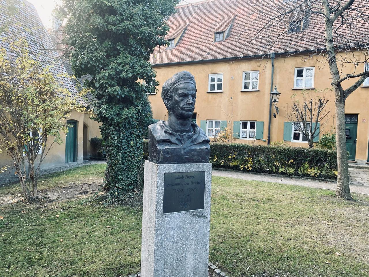 Auf einem Rasenstück steht eine bronzene Büste von Jakob Fugger, im Hintergrund sieht man die ockerfarbenen Häuser der Fuggerei. Auf einem Rasenstück steht eine bronzene Büste von Jakob Fugger, im Hintergrund sieht man die ockerfarbenen Häuser der Fuggerei.