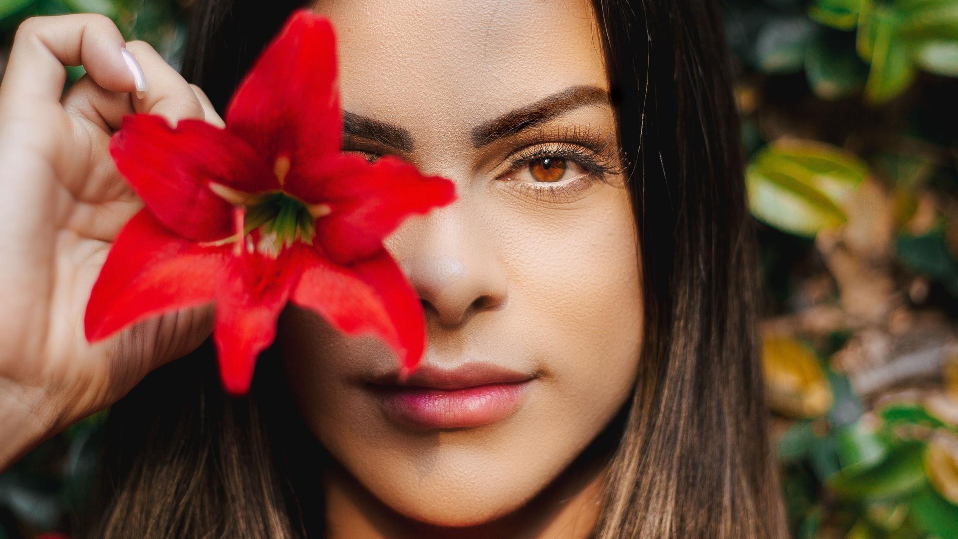 Eine junge, dunkelhaarige Frau hält sich vor ein Auge eine knallrote Blüte. Eine junge, dunkelhaarige Frau hält sich vor ein Auge eine knallrote Blüte.