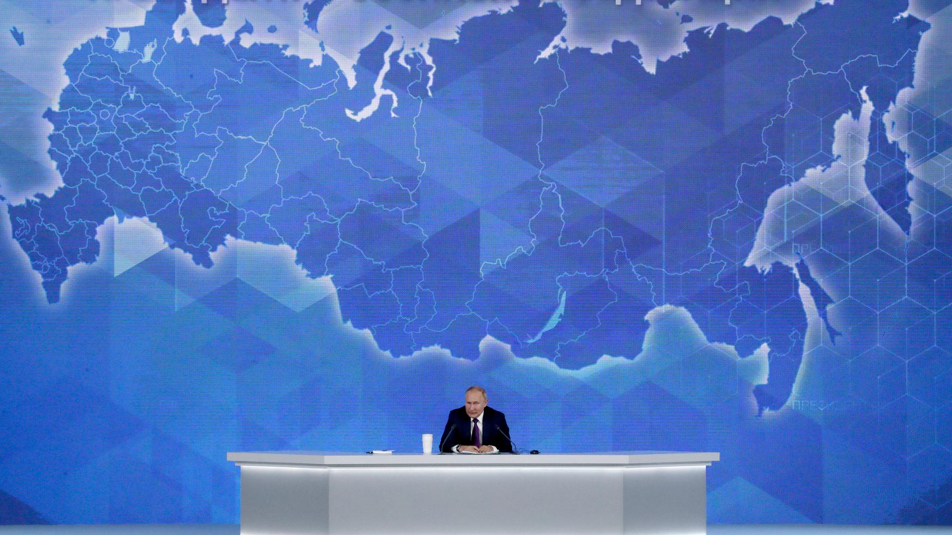 Wladimir Putin sitzt auf einem Podium mit einer rieseigen Russlandkarte im Hintergrund