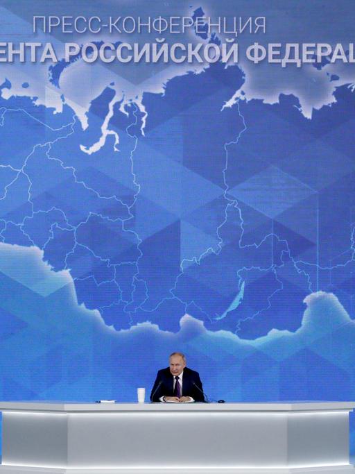Wladimir Putin sitzt auf einem Podium mit einer rieseigen Russlandkarte im Hintergrund