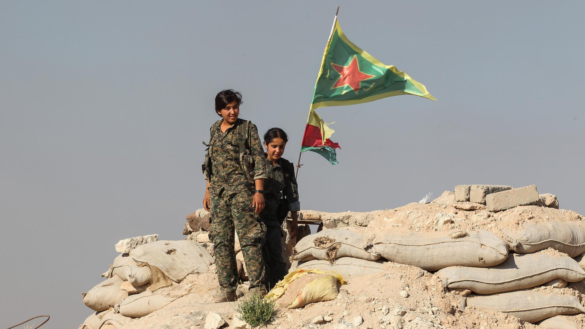 Zwei Frauen in Militäruniformen stehen auf einem Berg aus Schutt und Sandsäcken. Im Hintergrund weht die Flagge von Rojava und die der YPJ, eine Frauenverteidigungseinheit. Zwei Frauen in Militäruniformen stehen auf einem Berg aus Schutt und Sandsäcken. Im Hintergrund weht die Flagge von Rojava und die der YPJ, eine Frauenverteidigungseinheit.
