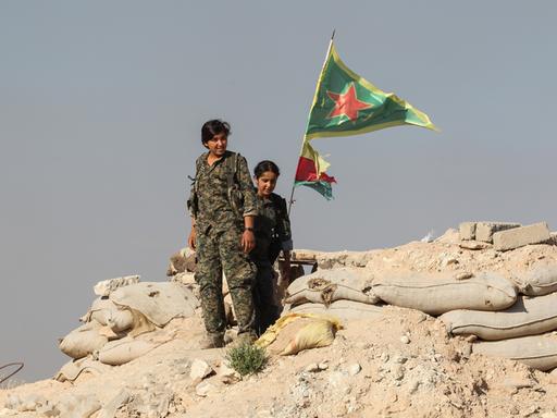 Zwei Frauen in Militäruniformen stehen auf einem Berg aus Schutt und Sandsäcken. Im Hintergrund weht die Flagge von Rojava und die der YPJ, eine Frauenverteidigungseinheit.