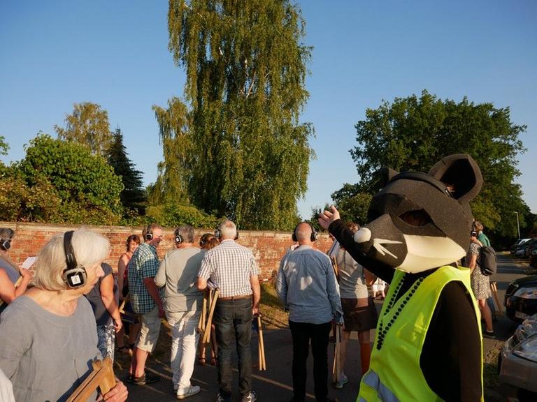 Theaterbesucher, die Kopfhörer tragen, begegnen im Freien einem Darsteller mit gelber Warnweste und einer Maus-Maske auf dem Kopf. Theaterbesucher, die Kopfhörer tragen, begegnen im Freien einem Darsteller mit gelber Warnweste und einer Maus-Maske auf dem Kopf.