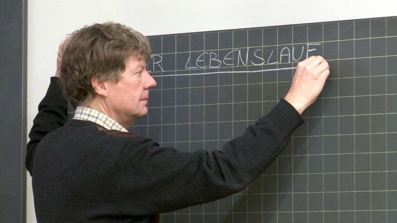 Christian Zingg steht vor einer Tafel und schreibt "Der Lebenslauf" mit Kreide. Christian Zingg steht vor einer Tafel und schreibt "Der Lebenslauf" mit Kreide.