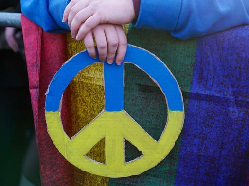 Zwei Hände halten das Symbol für Frieden in den ukrainischen Nationalfarben Blau und Gelb. Es handelt sich um eine Teilnehmende an einer Friedenskundgebung in Berlin anlässlich des Krieges in der Ukraine.