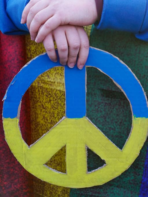Zwei Hände halten das Symbol für Frieden in den ukrainischen Nationalfarben Blau und Gelb. Es handelt sich um eine Teilnehmende an einer Friedenskundgebung in Berlin anlässlich des Krieges in der Ukraine.