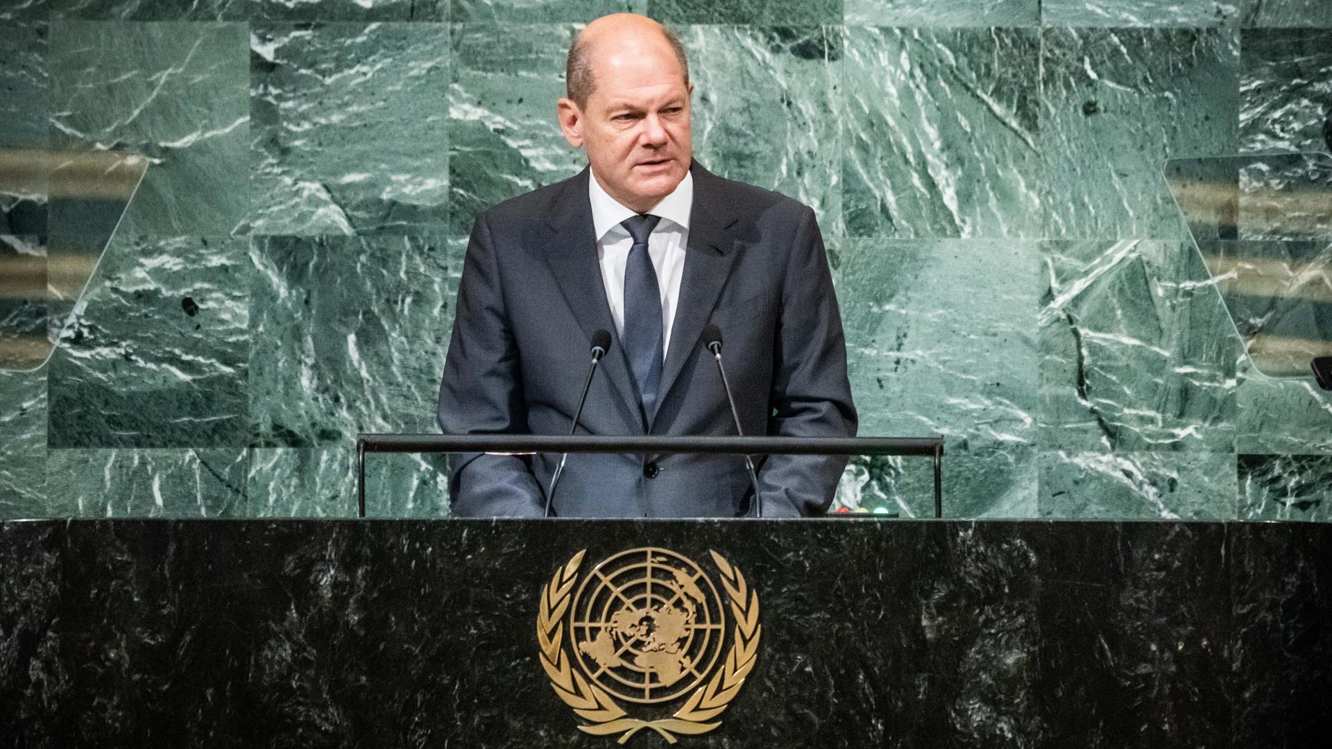 Bundeskanzler Olaf Scholz steht an einem Rednerpult, auf der Trennwand zum Publikum ist das goldene Wappen der Vereinten Nationen zu sehen. Bundeskanzler Olaf Scholz steht an einem Rednerpult, auf der Trennwand zum Publikum ist das goldene Wappen der Vereinten Nationen zu sehen.