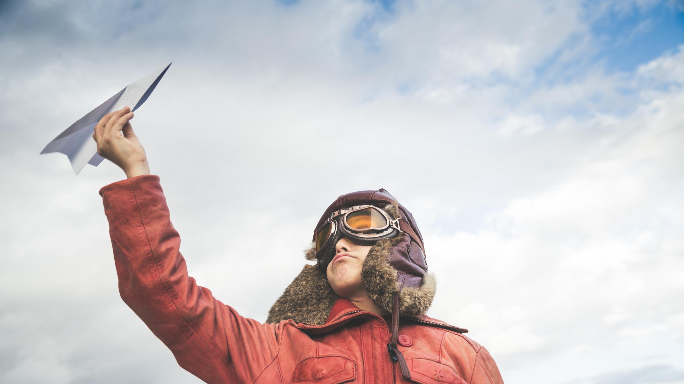 Ein Junge in Pilotenkostüm hält einen Papierflieger in die Luft. Ein Junge in Pilotenkostüm hält einen Papierflieger in die Luft.