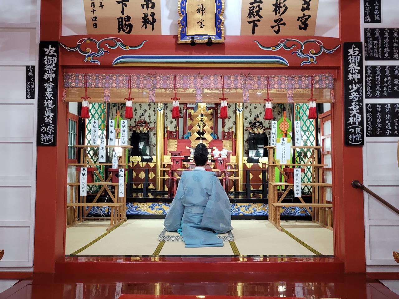 Priester Taishi Kato, in einem blauen Gewand, sitzt mit dem Rücken zu Kamera und wendet sich den Ornamenten und Figuren in der Nische des Shinto-Schreins zu.