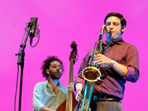 Ein Mann in braunem Hemd bläst Tenorsaxofon. Im Hintergrund spielt ein Mann Kontrabass. Der Bildhintergrund ist einheitlich pink. 