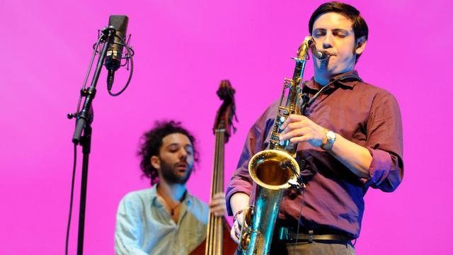 Ein Mann in braunem Hemd bläst Tenorsaxofon. Im Hintergrund spielt ein Mann Kontrabass. Der Bildhintergrund ist einheitlich pink. 