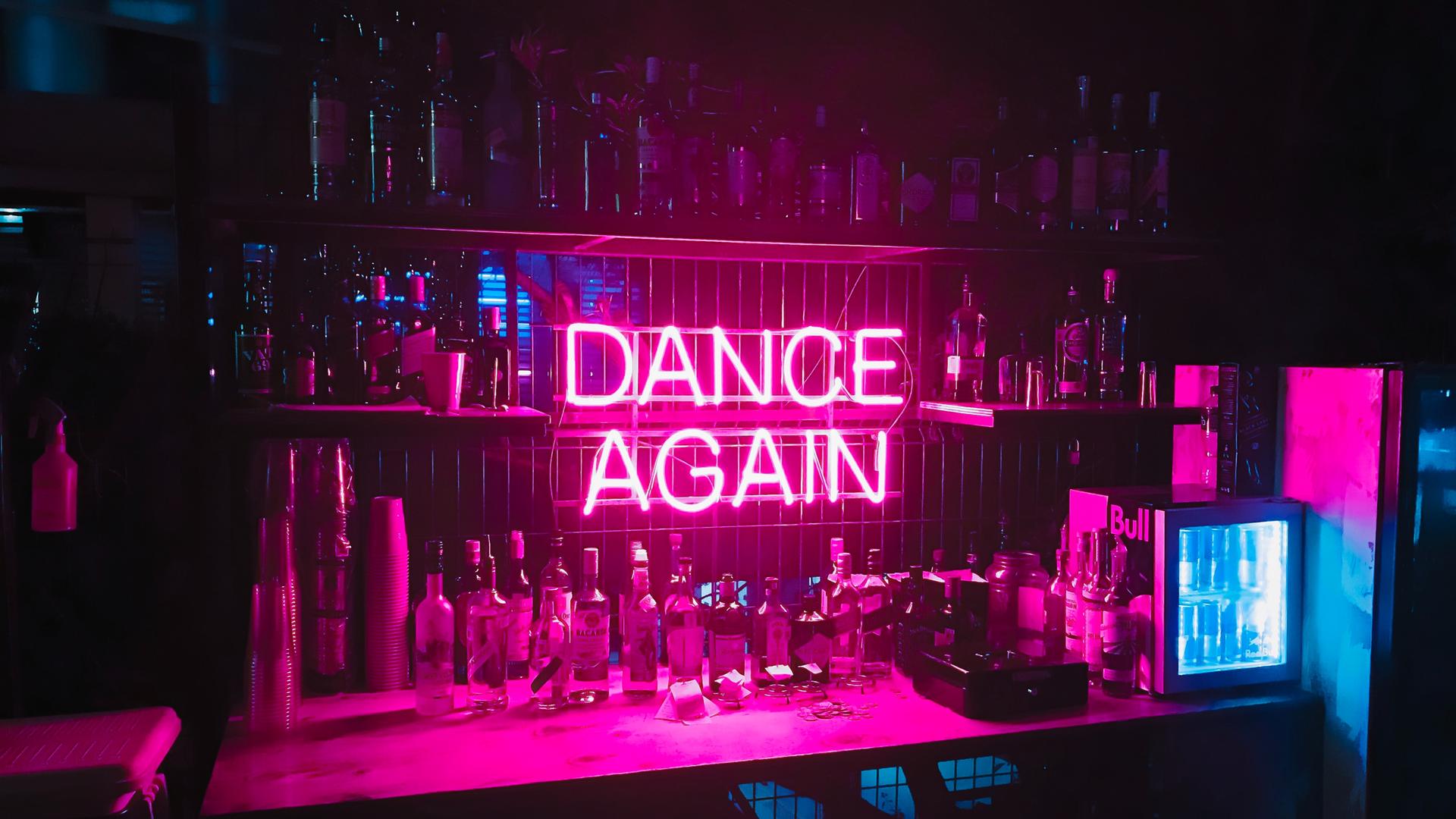 Hinter einer Bar ist in pinkfarbener Neonschrift der Schriftzug "DANCE AGAIN" angebracht. Hinter einer Bar ist in pinkfarbener Neonschrift der Schriftzug "DANCE AGAIN" angebracht.